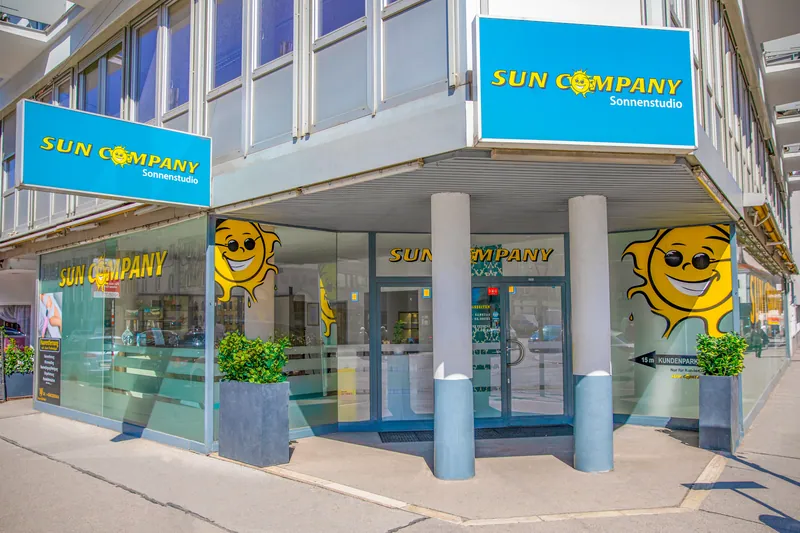Ugaona zgrada sa plavim Sun Company Sonnenstudio tablama, staklenom fasadom, nasmejanim suncima i žardinjerama ispred ulaza