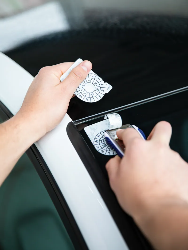 Ruke strugačem skidaju staru kontrolnu nalepnicu sa stakla automobila, dok drže novu nalepnicu