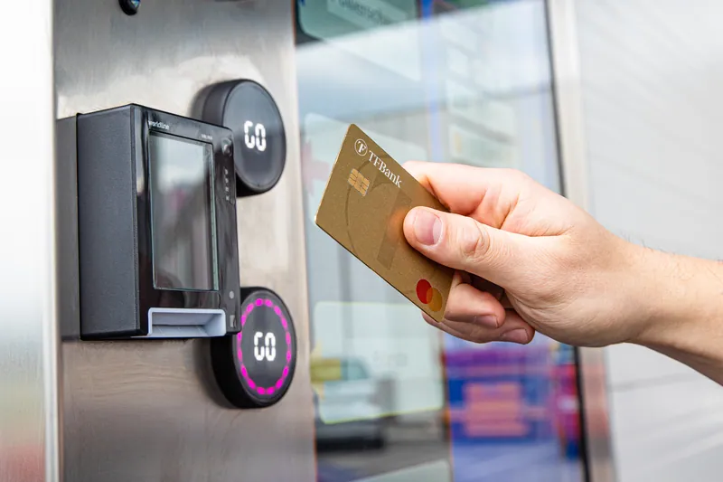 Ruka drži zlatnu TF Bank Mastercard kreditnu karticu ispred Worldline čitača pored osvetljenih Go dugmadi na terminalu perionice