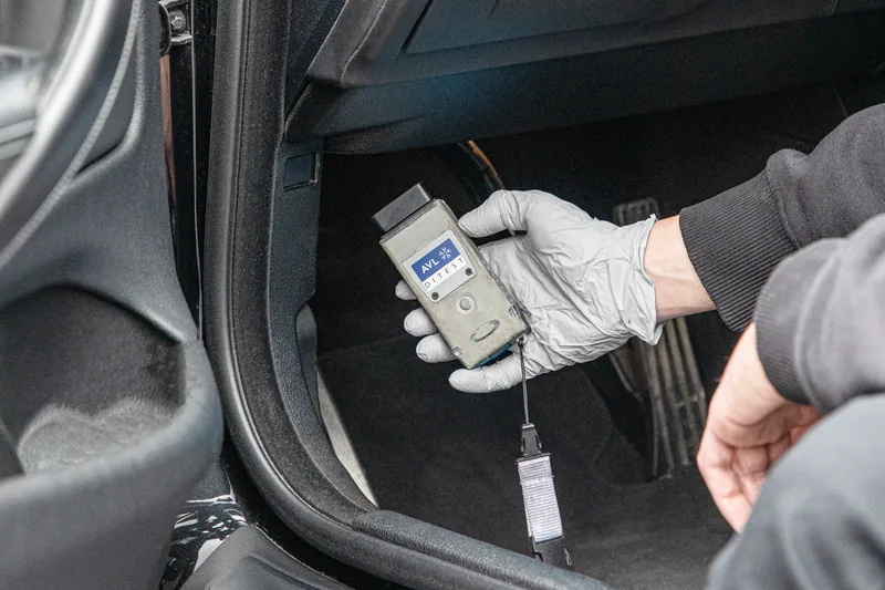 Ruka u rukavici drži AVL DiTEST dijagnostički uređaj u prostoru za noge u automobilu, pedale se vide iza
