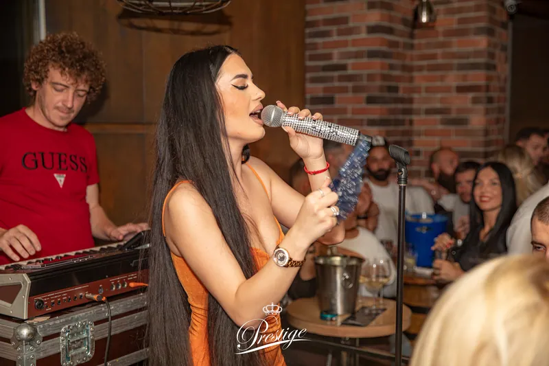 Pevačica duge crne kose peva u mikrofon, pored klavijaturista u Guess majici, Prestige