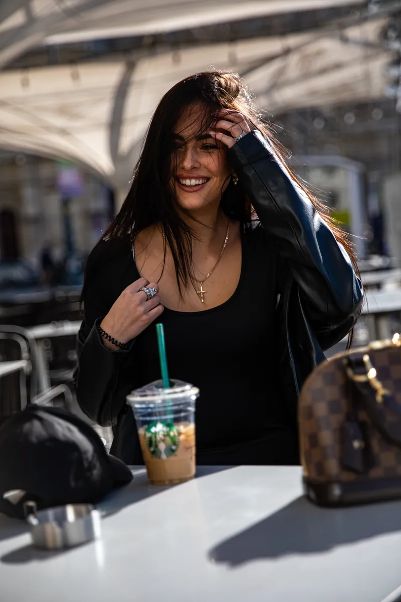 Nasmejana žena za belim stolom sa Starbucks ledenom kafom, crnom kapom i torbom