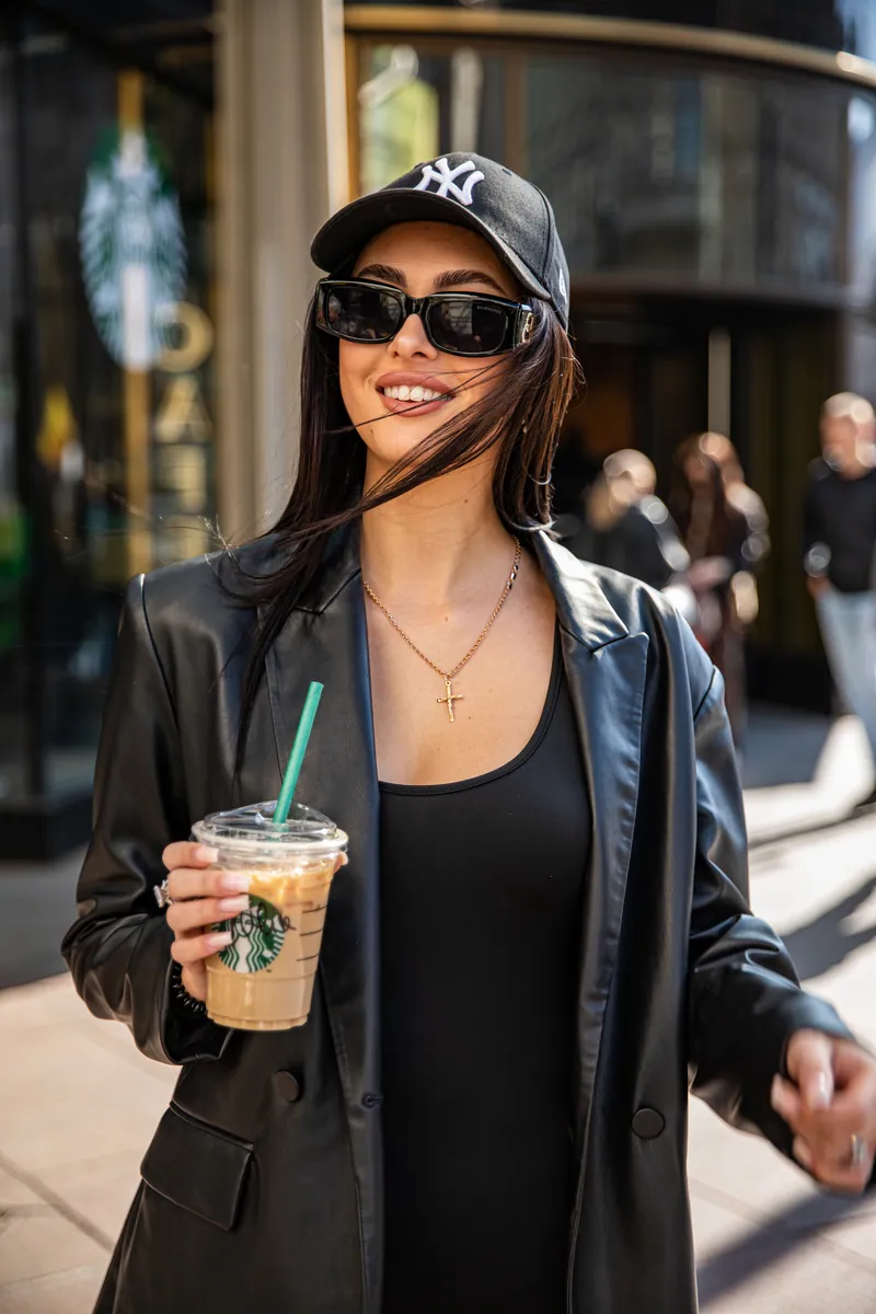 Nasmejana žena u kapi, sa naočarima i kožnim sakoom drži Starbucks ledenu kafu